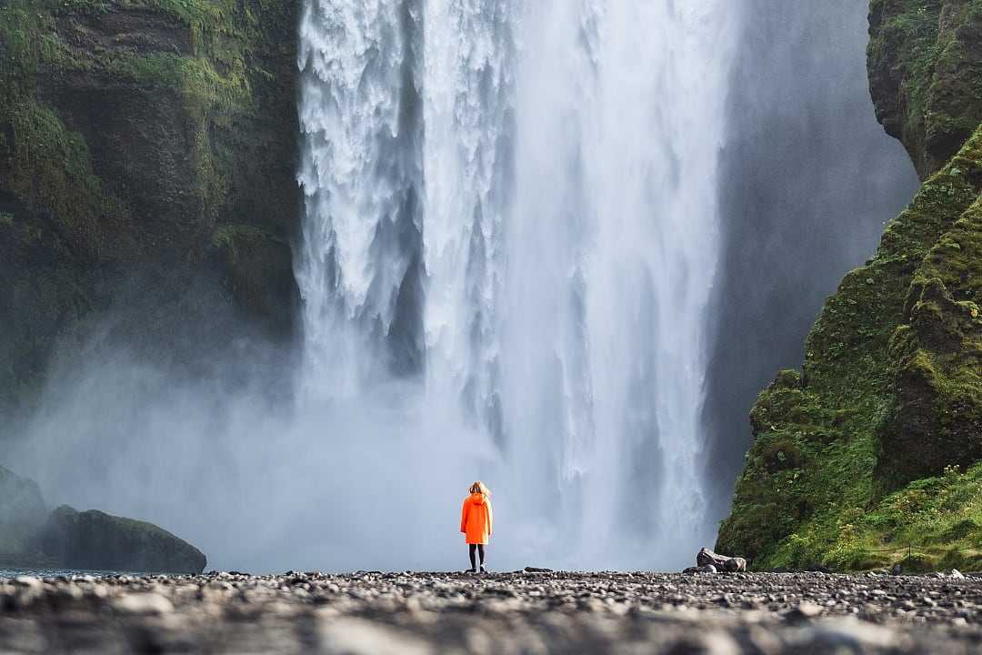 Woman in orange jacket standing at the base of Skogafoss Waterfall in Iceland