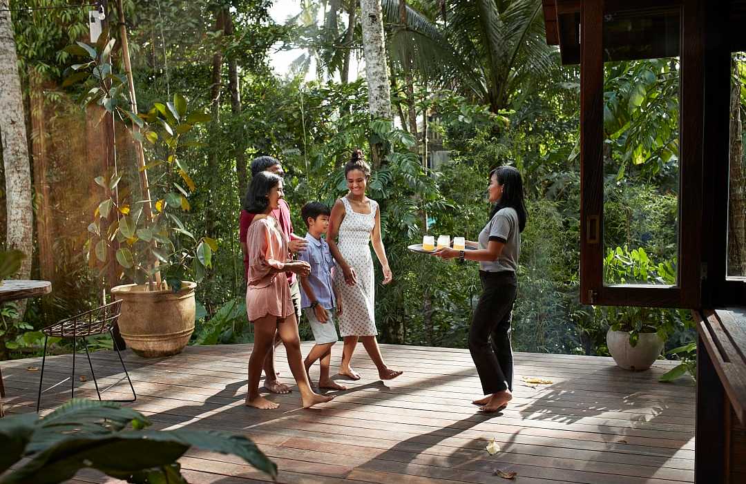 A female specialist stands on a sunny tropical balcony, smiling and gesturing while speaking to a traveling family.