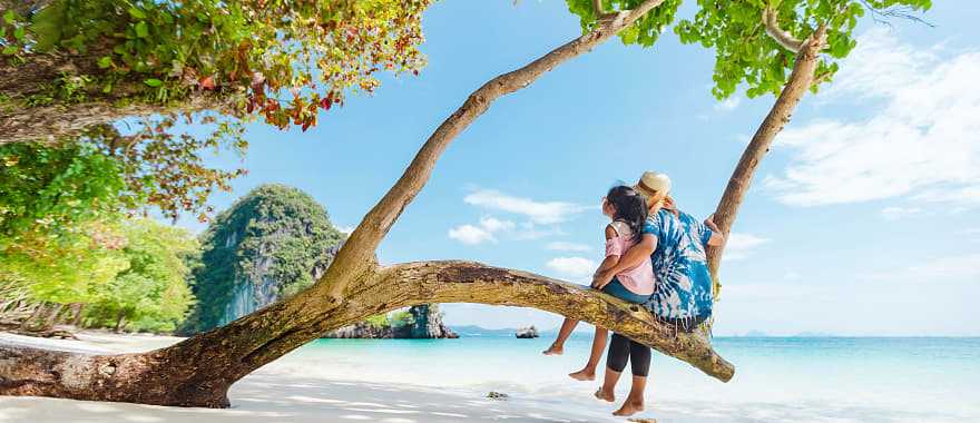 Mother and daughter sitting on a low tree branch on a beach in Phuket, Thailand
