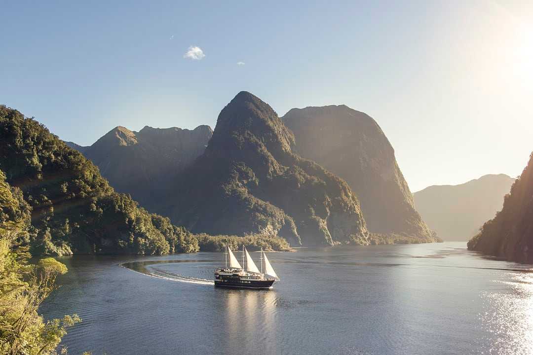 Sailing vessel cruises through tranquil fjord beneath towering forested peaks.