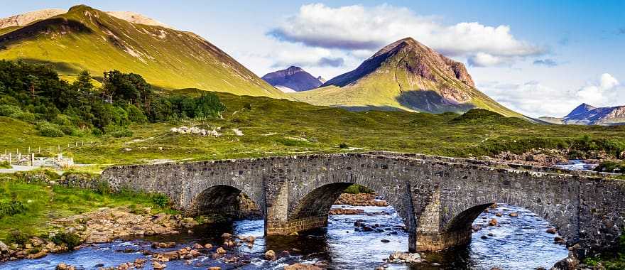 Old vintage brick bridge crossing river in Sligachan, Isle of Skye, Scotland with blue sky, hills and mountains in the background