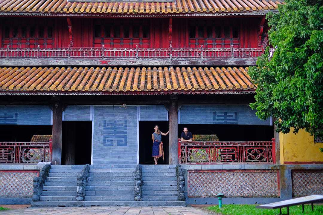Travelers at the Imperial City of Hue, Vietnam, standing outside a historic temple with red columns and tiled roof.