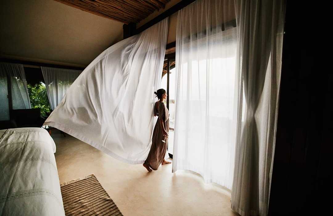A woman in a flowing dress stands by large windows as a sheer white curtain billows in a luxury hotel room.