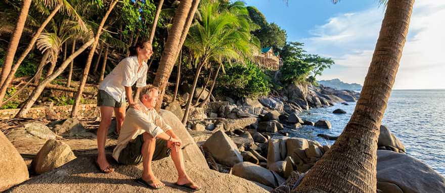 Phuket, Thailand Senior couple enjoying a tropical beach at sunset surrounded by palm trees and rocky shoreline in Thailand.