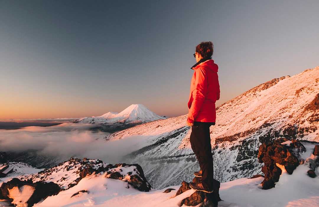 Hiker in winter jacket standing on snowy mountain, admiring view of snow-capped peaks and clouds at sunrise in Tongariro National Park, New Zealand