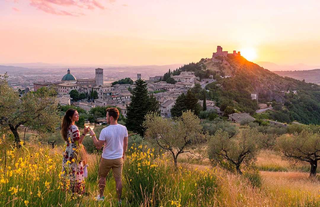 Umbria, Italy Couple toasting with wine at sunset overlooking a picturesque medieval town surrounded by olive trees and rolling hills