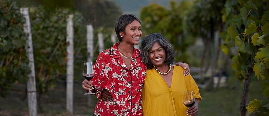 Smiling mother and daughter holding wineglasses and standing with arms around each other at a vineyard