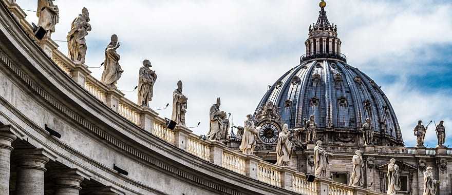 Intricate statues standing on a rooftop with the peak of St Peter's Basilica in the background.