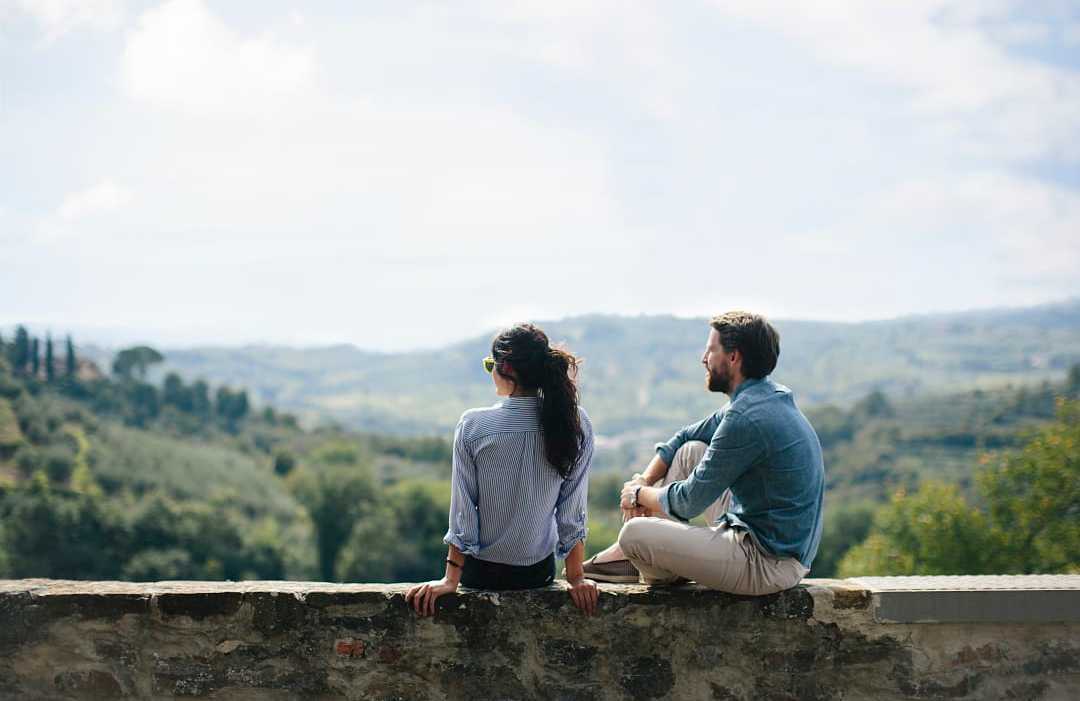 Couple sitting on stone wall overlooking rolling countryside hills in Tuscany, Italy